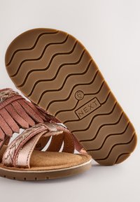 Sandali color oro rosa con cinturini intrecciati, finitura in suede, soletta imbottita e suola in gomma marrone testurizzata con motivo ondulato.