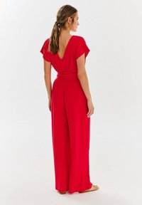 Czerwony kombinezon z dekoltem w kształcie V, krótkimi rękawkami, szerokimi nogawkami i paskiem w talii. Gładki materiał o zwiewnej fakturze.