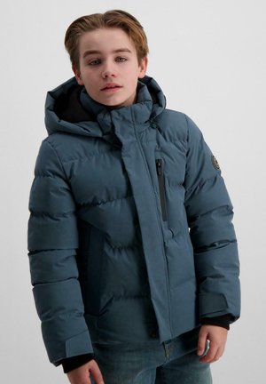 Een jonge persoon met blond haar draagt een blauwe gewatteerde jas en jeans, poseert tegen een effen witte achtergrond.
