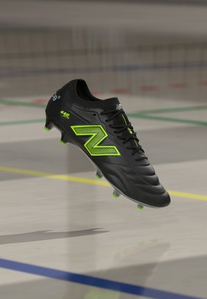 Črne nogometne kopačke New Balance z neonsko zelenimi akcenti in študijem, ki lebdi nad talno površino notranjega športnega igrišča.