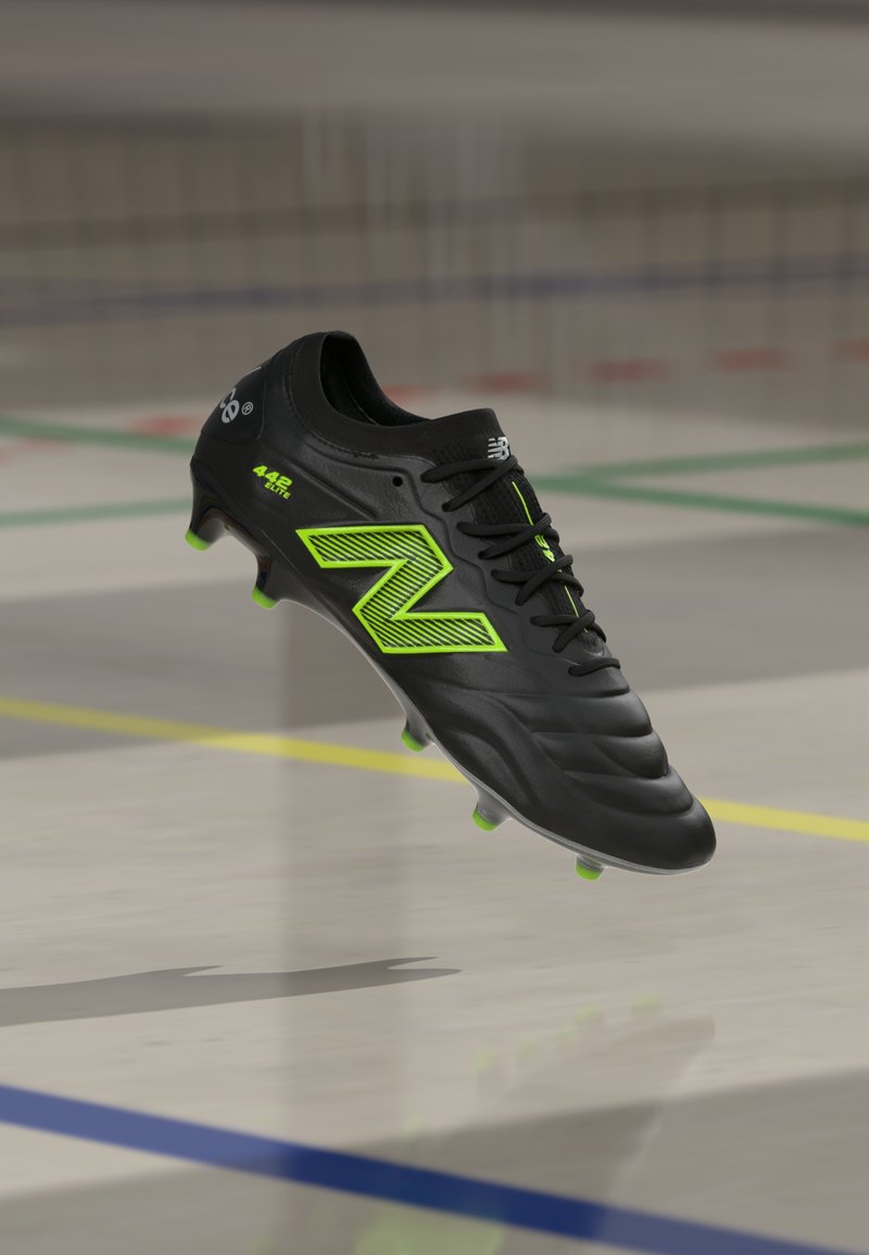 Juodas „New Balance“ futbolo batelis su neoninės žalios spalvos akcentais ir taškais, plūduriuojantis virš vidaus sporto aikštelės grindų.