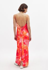 Moteris su trumpais rudais plaukais, apsirengusi be rankovių, atvira nugara ilga suknele su oranžiniais ir rožiniais gėlių raštais bei baltais batais, stovi ant baltų grindų.