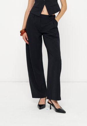Femme portant un pantalon noir à jambes larges, un gilet noir sans manches, des talons slingback noirs à bout pointu et des bracelets marron épais au poignet.