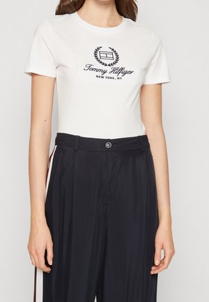 Kobieta ubrana w białą koszulkę Tommy Hilfiger z logo oraz czarne, wysokowodne spodnie z bocznymi paskami.