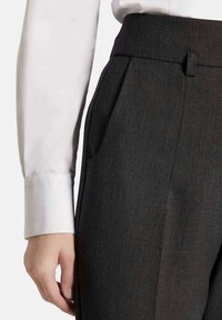 Donkergrijze op maat gemaakte broek van gestructureerde stof, met voorplooien, zijzakken en een gladde tailleband. Witte overhemdmanchet zichtbaar.