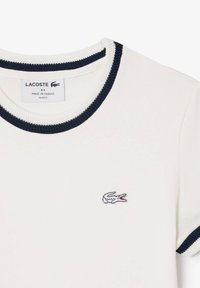 Camiseta blanca de corte ajustado con cuello y puños de canalé en azul marino, que presenta un pequeño logo de cocodrilo bordado en el pecho. Fabricada en Francia.