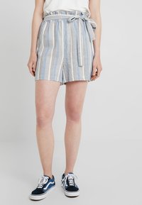 Frau trägt hoch geschnittene Shorts in Blau und Weiß mit vertikalen Streifen und einem in eine Schleife gebundenen Gürtel, ein weißes Oberteil und blaue Sneakers mit weißen Schnürsenkeln.