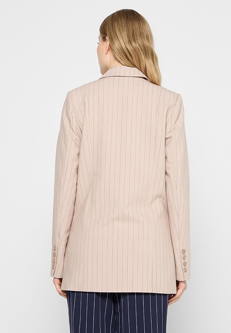 Femme aux longs cheveux blonds portant un blazer beige à rayures et un pantalon bleu marine à rayures, vue de dos contre un fond uni.