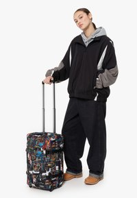 Czarna walizka na kółkach z kolorowym graffiti, materiał tekstylny, podwójne uchwyty i wysuwany metalowy stelaż. Duży i prostokątny kształt.