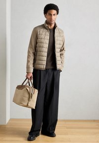 Veste matelassée beige clair avec un design texturé, associée à un pull en tricot marron foncé et un pantalon noir large. Tient un sac de voyage beige clair.