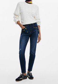 Valkoinen palmikkoneule, tummansiniset farkut, joissa on kukkakirjontaa ja kuluneita kohtia. Mustat kuviolliset lenkkarit täydentävät asukokonaisuuden.