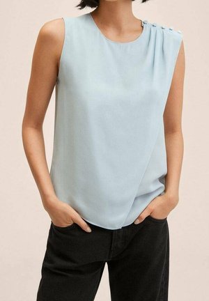 Bezrękawowa jasnoniebieska bluzka z guzikami na lewym ramieniu, noszona z czarnymi spodniami, ręce w kieszeniach.