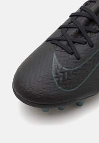 Black football boot with textured synthetic upper, featuring black laces and a subtle green Nike logo. Rounded toe, molded studs on the sole.