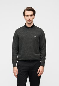 Νέος άνδρας φορώντας σκούρο γκρι πουλόβερ με στρογγυλή λαιμόκοψη πάνω από μαύρο πουκάμισο με γιακά και μαύρο παντελόνι, στέκεται μπροστά σε απλό ανοιχτόχρωμο φόντο.