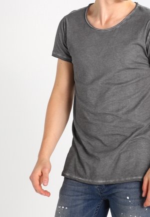 Person iført en kortærmet, falmet grå T-shirt og blå jeans med hvide malingstænk, stående mod en ensartet lys baggrund.