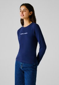 Mlada ženska v temno modri majici z dolgimi rokavi z napisom "RED BULL RACING", oblečena v modre kavbojke, stoji na sivem ozadju.