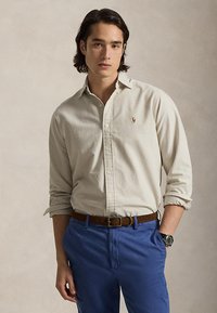 Junger Mann mit beiger Hemdbluse, blauer Hose, braunem Gürtel und Armbanduhr, steht mit einer Hand in der Tasche vor neutralem Hintergrund.