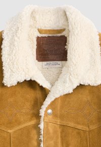 Tan suède jack met een crèmekleurige nepbont kraag, met decoratieve stiksels en een leren label aan de binnenkant. Zilveren drukknoopjes zichtbaar.