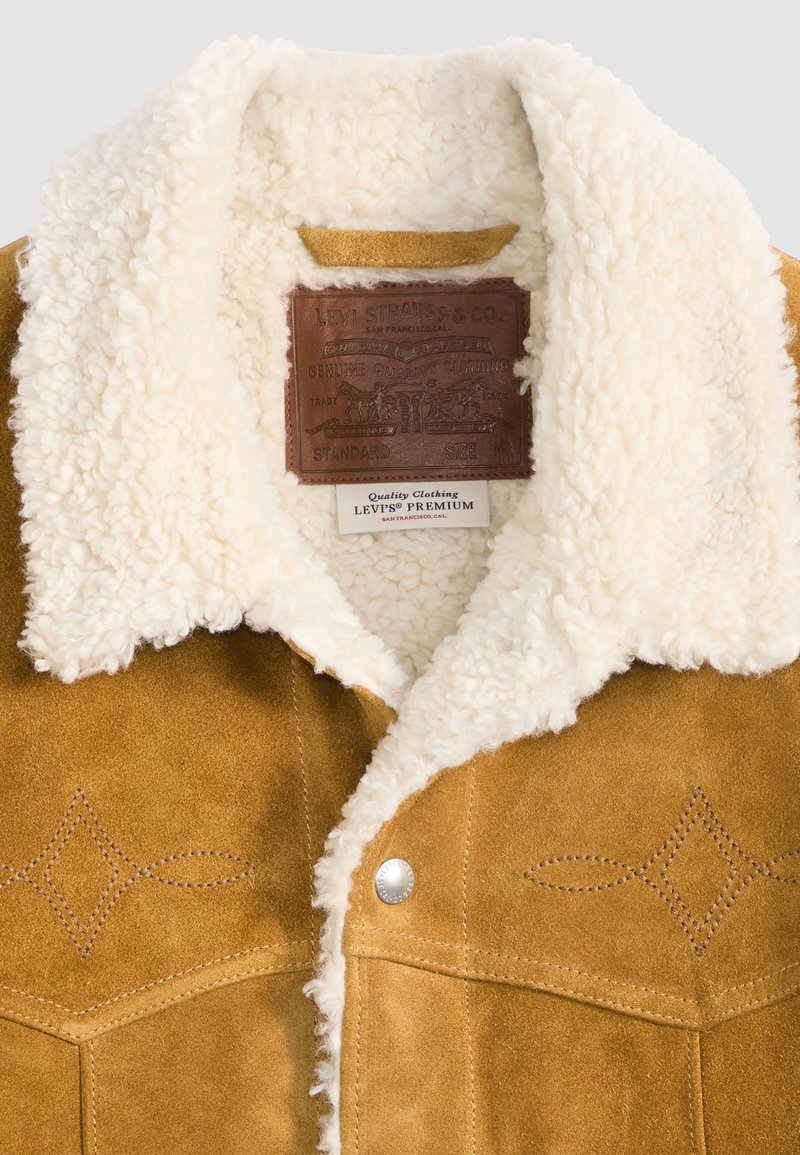 Tan suède jack met een crèmekleurige nepbont kraag, met decoratieve stiksels en een leren label aan de binnenkant. Zilveren drukknoopjes zichtbaar.