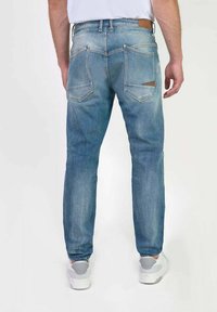 Jean en denim bleu clair avec une coupe fuselée, comportant deux poches arrière, des coutures contrastées et une étiquette en cuir marron sur la ceinture.