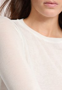 Top leggero e trasparente di colore bianco con una texture morbida e liscia. Presenta un collo rotondo e maniche lunghe con dettagli di cucitura minimi.