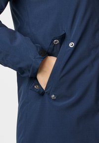 Granatowa kurtka o gładkiej, matowej teksturze, z boczną kieszenią i zapięciem na napy. Akcenty obejmują srebrne elementy metalowe oraz rzep.