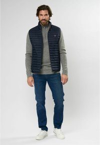 Gilet trapuntato navy sopra un maglione grigio chiaro, abbinato a jeans blu e scarpe da ginnastica bianche. Tessuti morbidi, zip frontale, senza loghi visibili sui jeans.
