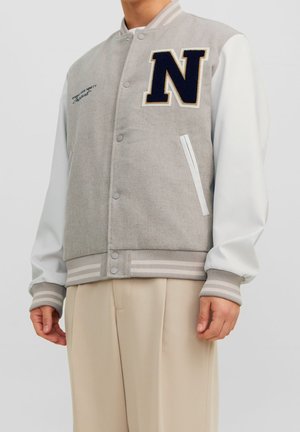 Mężczyzna ubrany w jasnoszarą kurtkę varsity z białymi rękawami, granatową łatą z literą "N" na piersi oraz beżowe spodnie, stojący na jednolitym tle.