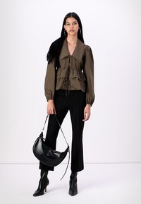 Blouse péplum vert olive avec col et cordon de serrage, associée à un pantalon noir ajusté et un sac à main noir, sur fond blanc.
