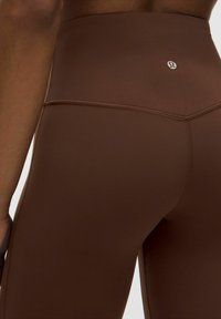 Brązowe legginsy z wysokim stanem, wykonane z elastycznego materiału, z płaskimi szwami i małym logo na tylnej części pasa.