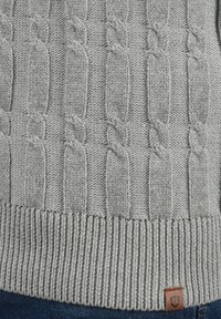Pull en maille torsadée gris avec des motifs texturés et un ourlet côtelé. Comprend une petite étiquette en cuir au bas.