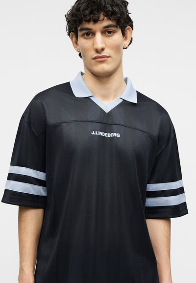 Jeune homme aux cheveux courts et bouclés portant un maillot de sport noir avec un col et des rayures sur les manches bleu clair, logo "J. Lindeberg" sur la poitrine.