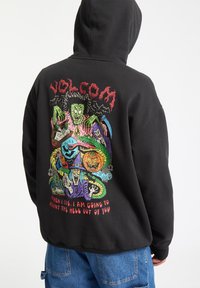 Zwarte hoodie met een kleurrafiek op de achterkant met gestileerde figuren, waaronder een heks, pompoen en slang, met tekst eronder.