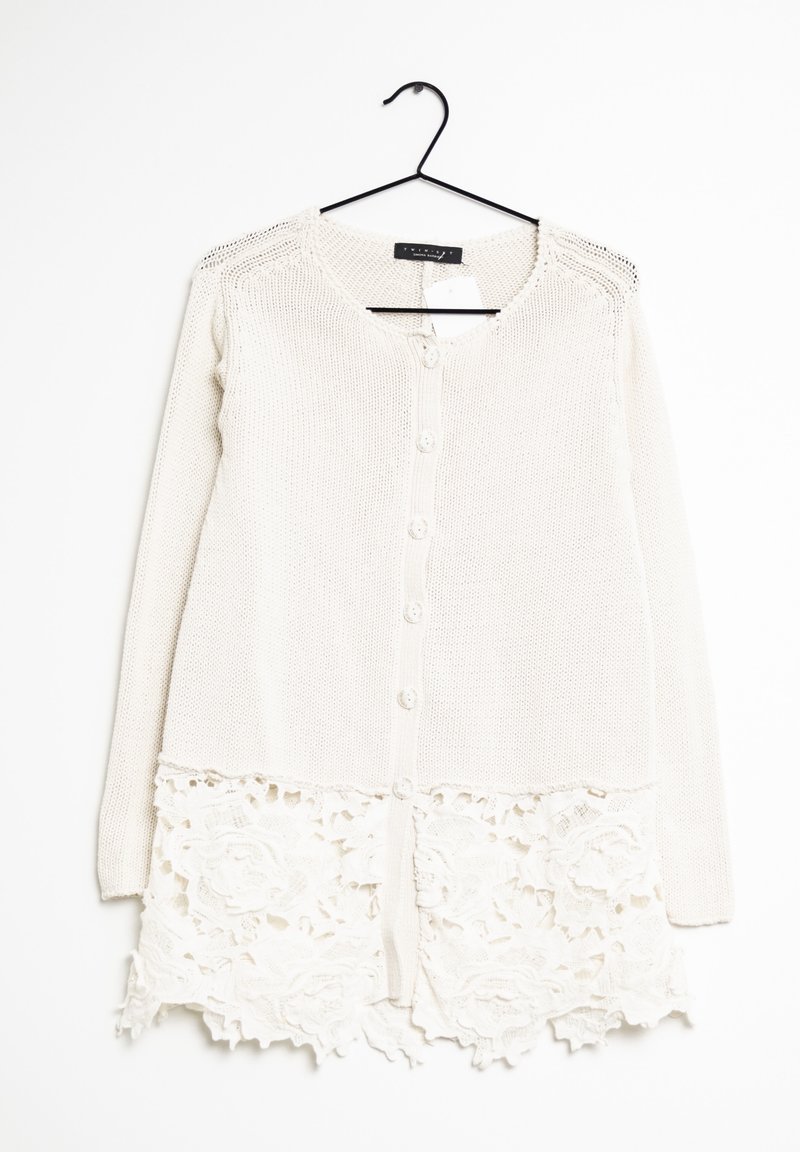 Valkoinen neuletakki, jossa on teksturoidusta neuleesta valmistettu runko ja pitsireuna helmassa, pitkähihaisena ja seitsemällä nappilla edessä.