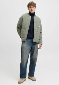 Veste matelassée vert olive avec fermeture à pression à l'avant et poche à patch, portée sur un col roulé bleu marine. Assortie à un jean bleu délavé et des chaussures claires.