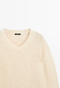 Pull en maille couleur crème avec un décolleté en V et un col côtelé. Coupe décontractée avec un tissu texturé et sans motifs ni embellissements visibles.