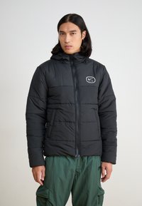 Black padded jacket with a zip front and adjustable hood. Features a white Nike Air logo on the chest. Worn with green cargo pants.