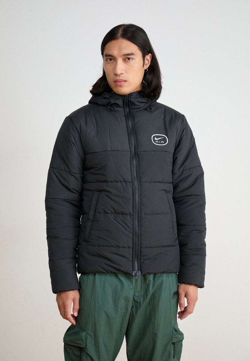 Black padded jacket with a zip front and adjustable hood. Features a white Nike Air logo on the chest. Worn with green cargo pants.