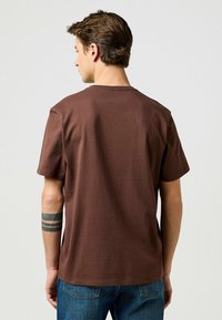 Ruskea puuvillapaita, jossa on pyöreä kaula-aukko ja lyhyet hihat. Paidassa on rento istuvuus ja tasainen pinta, nähtynä takaa.