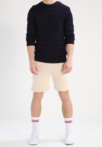Pull en maille bleu marine avec des rayures horizontales et des boutons aux épaules, porté avec un short beige, des baskets blanches et des chaussettes blanches à rayures rouges.
