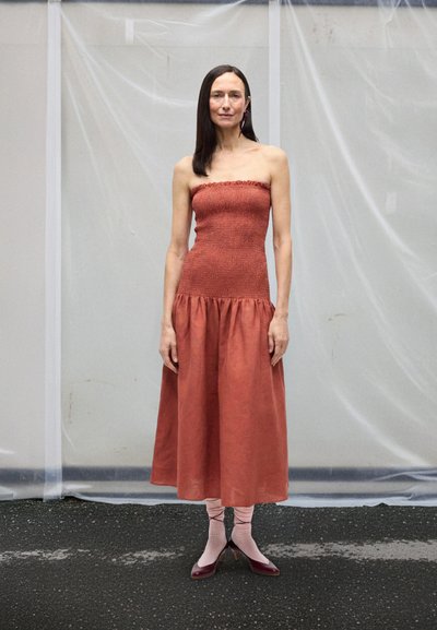 Sieviete ar gariem tumšiem matiem valkā bezplecu rūsu krāsas kleitu, rozā zeķes un bordo augstpapēžu kurpes, stāvot pret gaišu, strukturētu fonu.