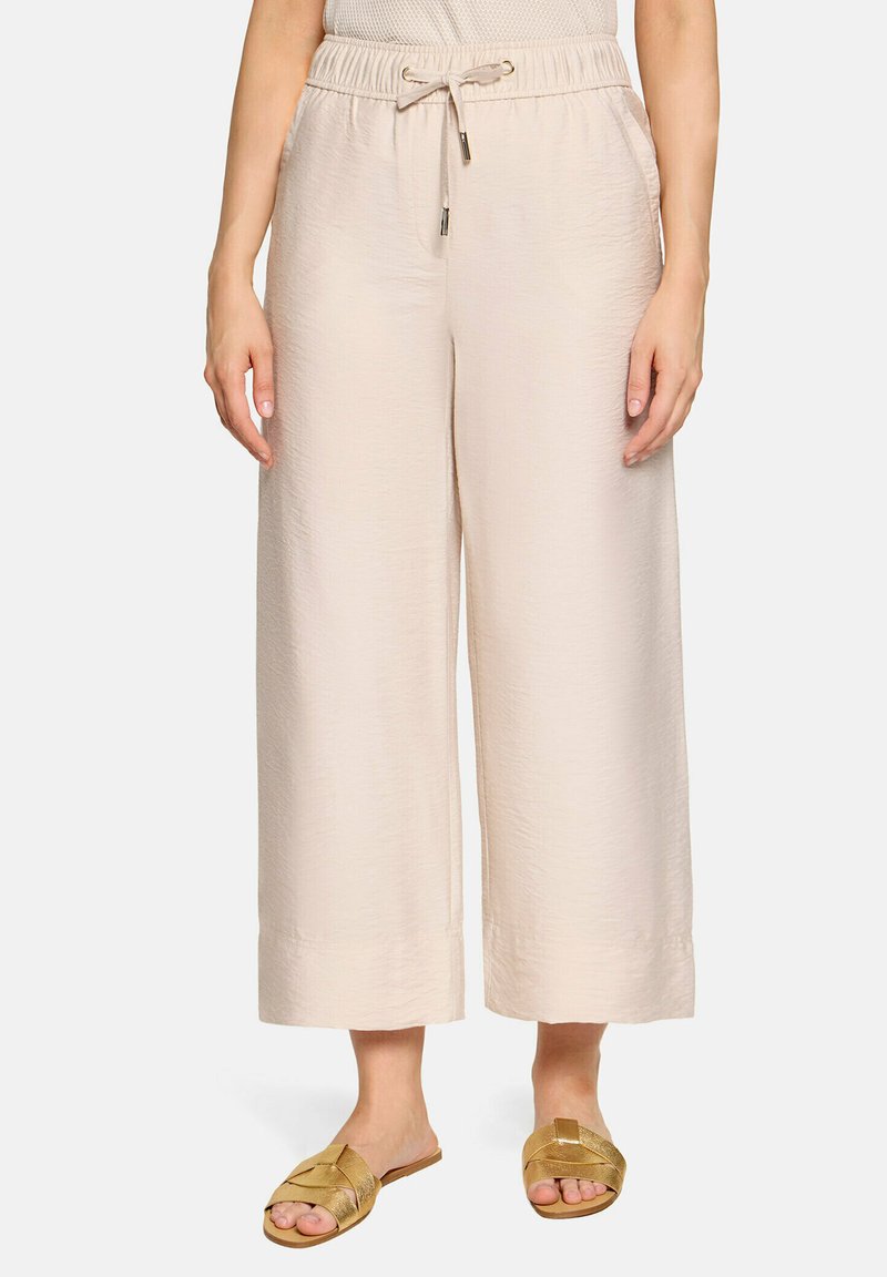 Femme portant un pantalon beige clair large et court avec taille à cordon et des sandales dorées, debout sur fond blanc.