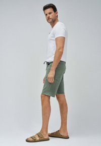 Pantalones cortos de algodón verde con bolsillos laterales, combinados con una camiseta blanca ajustada y sandalias marrones con doble hebilla. Diseño sencillo, ajuste casual.