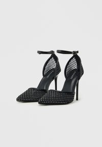 Zapatos de tacón alto negros con punta afilada, paneles laterales de malla y adornos de tachuelas en la parte delantera, que cuentan con una correa en el tobillo.
