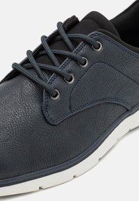 Mörkblå lädersko med texturerad yta, rund tå och kontrasterande vit gummisula. Har grå snören och metalliska öglor.