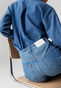 Veste en denim bleu moyen avec une coupe ample et des plis dans le dos. Le jean bleu comporte une étiquette logo blanche au niveau de la taille arrière.