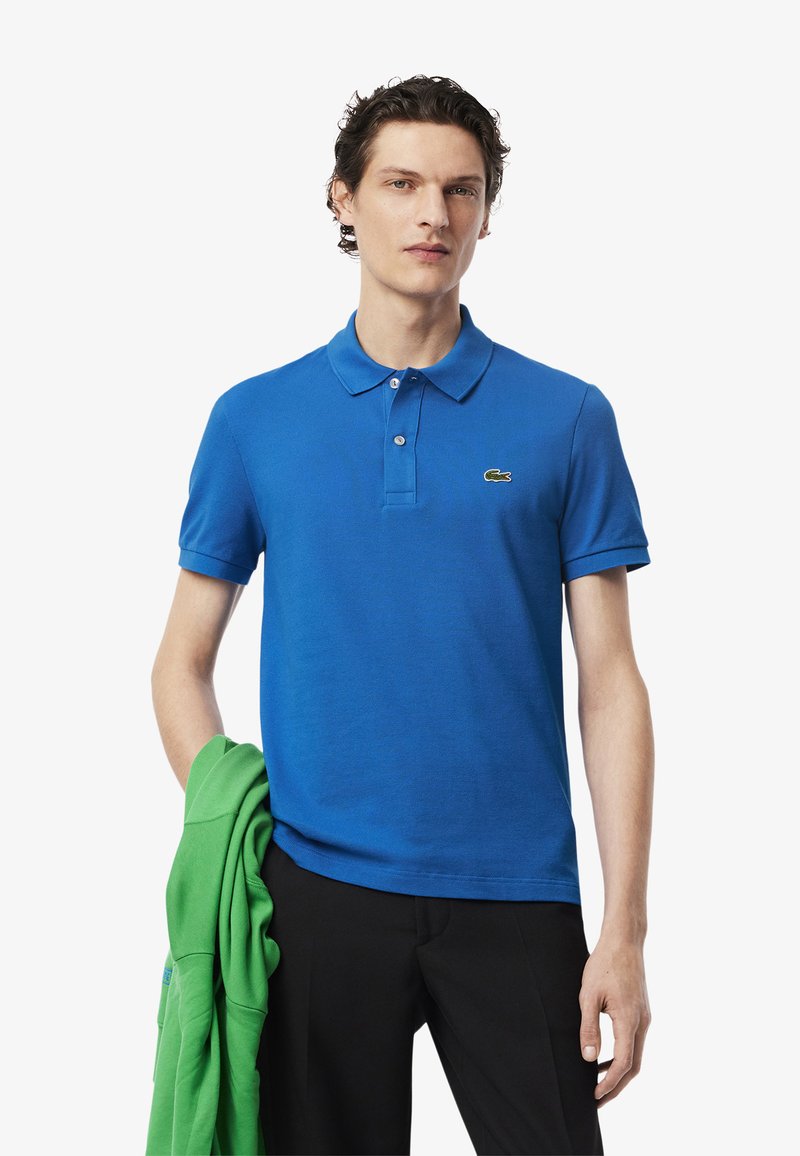 Camisa polo azul de algodón, con cuello, tres botones y un pequeño logo verde en el pecho. Combinada con un sudadera verde.