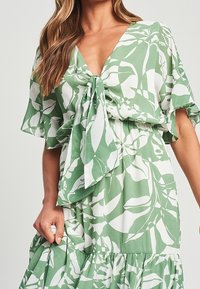 Vestido verde con patrón de hojas blancas, presenta un escote en V, detalle de nudo en la parte frontal y mangas de volantes. Tela suave con dobladillo en capas.