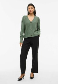 Cardigan côtelé vert clair avec un col en V, cinq boutons marron et des manches longues, associé à un pantalon slim noir et des mules à talons noires.