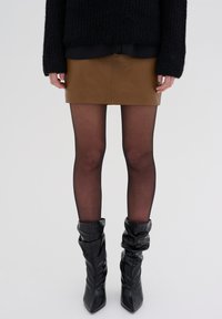 Mini gonna in pelle marrone con un design sartoriale, abbinata a delle calze sheer nere e stivali neri alti e morbidi con punta affilata.